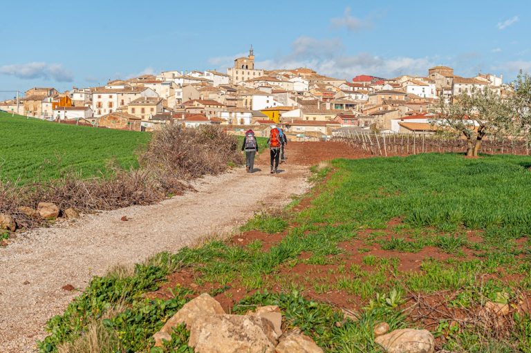 Two pilgrims with backpacks walk towards the picturesque village of Cirauqui, Spain, on a sunny day.